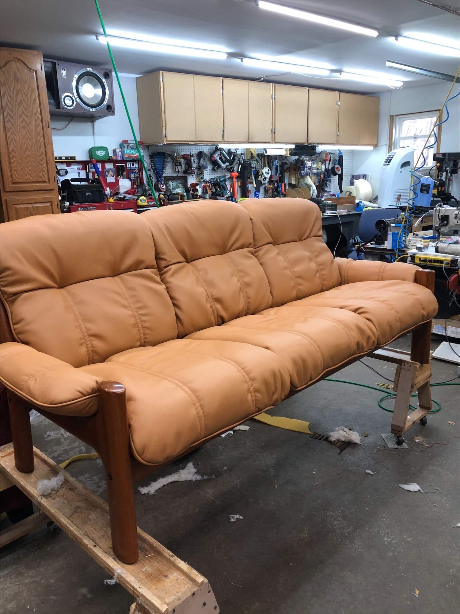 A light brown faux leather couch with tufted upholstery is shown in a cluttered workshop, surrounded by tools and equipment.
