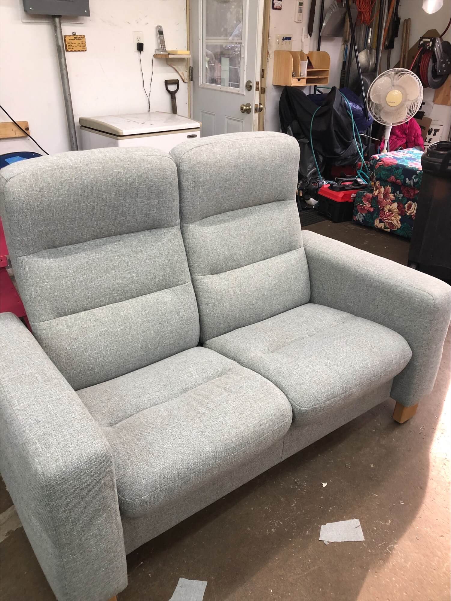 Light gray upholstered loveseat with a simple, modern design, placed in a garage setting filled with tools and equipment.